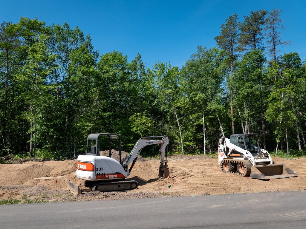 CROW WING CO, MN - 6 JUN 2021: A Bobcat mini excavator and front loader, with skid steer tracks, sits on dirt at a tree lined empty building lot, in preparation for new home construction. Excavation Services Southington CT